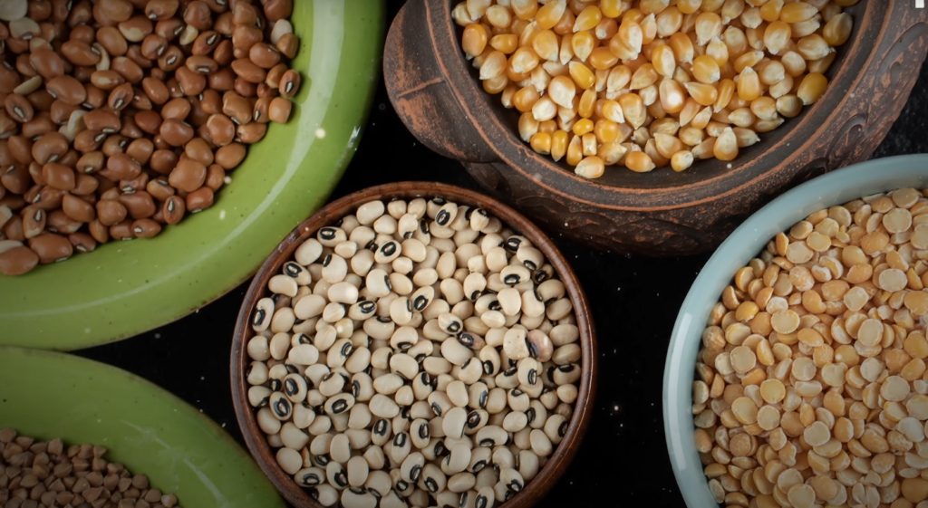 Millets in Hindi: विभिन्न प्रकार की मिलेट्स और अनाज कटोरों में दिखाए गए हैं, जिनमें मकई, काली आंख वाली लोबिया, अरहर की दाल और अन्य देसी अनाज शामिल हैं। यह छवि भारत में पारंपरिक पौष्टिक अनाजों के विविधता को दर्शाती है।